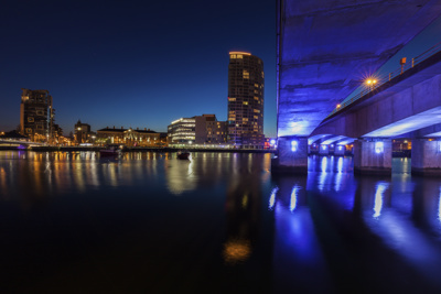 A night-time cityscape of Belfast, Ireland, featuring a lit bridge with blue lights reflecting on the calm water. A tall modern building and surrounding structures are illuminated in the background against a dark blue sky.