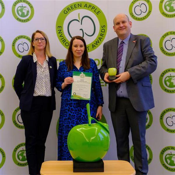 Three people standing in front of a Green Apple Environment Awards backdrop, holding a certificate and a trophy. In the foreground, a large green apple trophy is displayed on a table. The individuals are dressed formally, smiling at the camera.